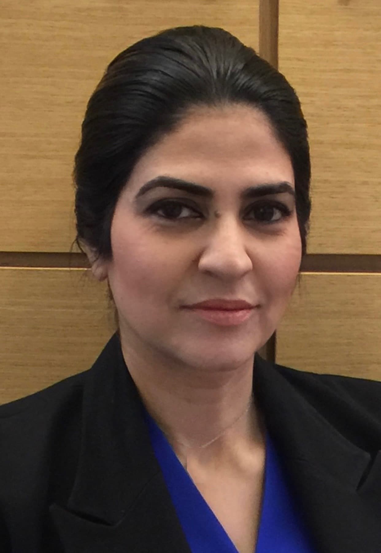 Woman with dark hair wearing a black blazer and blue top against wood paneling.