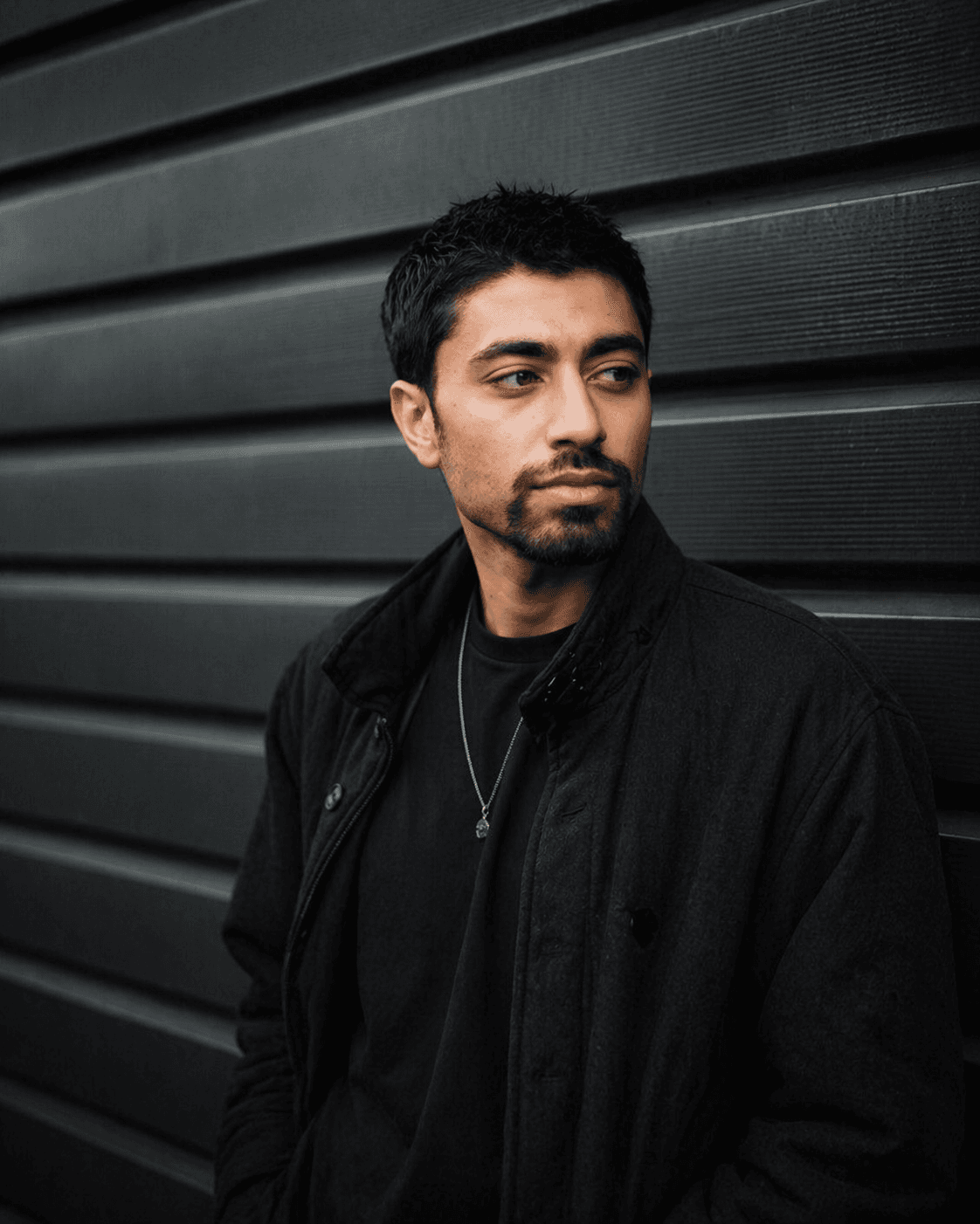 Man with dark hair and goatee wearing a black jacket against a slatted wall.