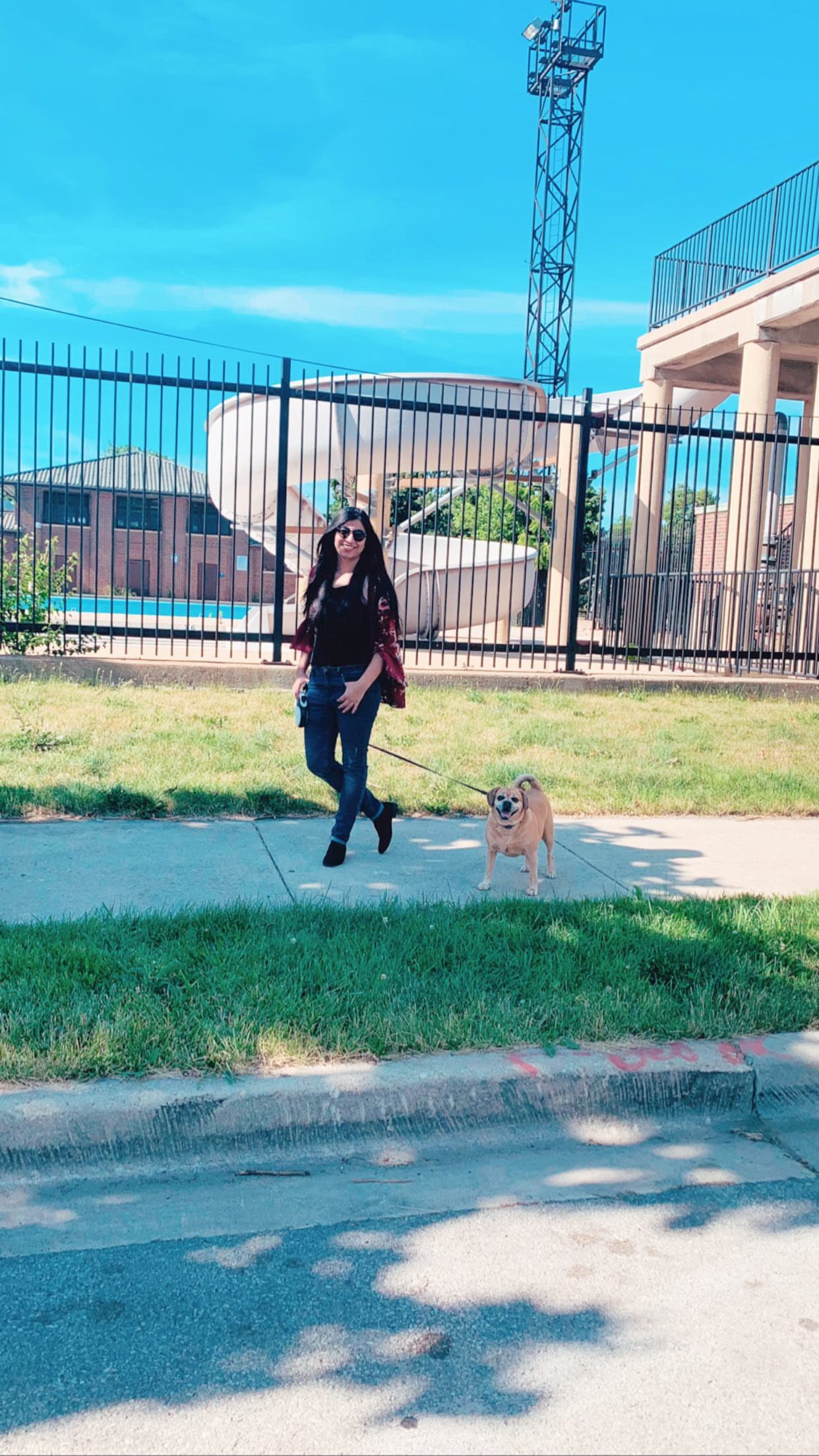 Smiling woman in maroon cardigan walks her small tan dog on a sunny sidewalk.