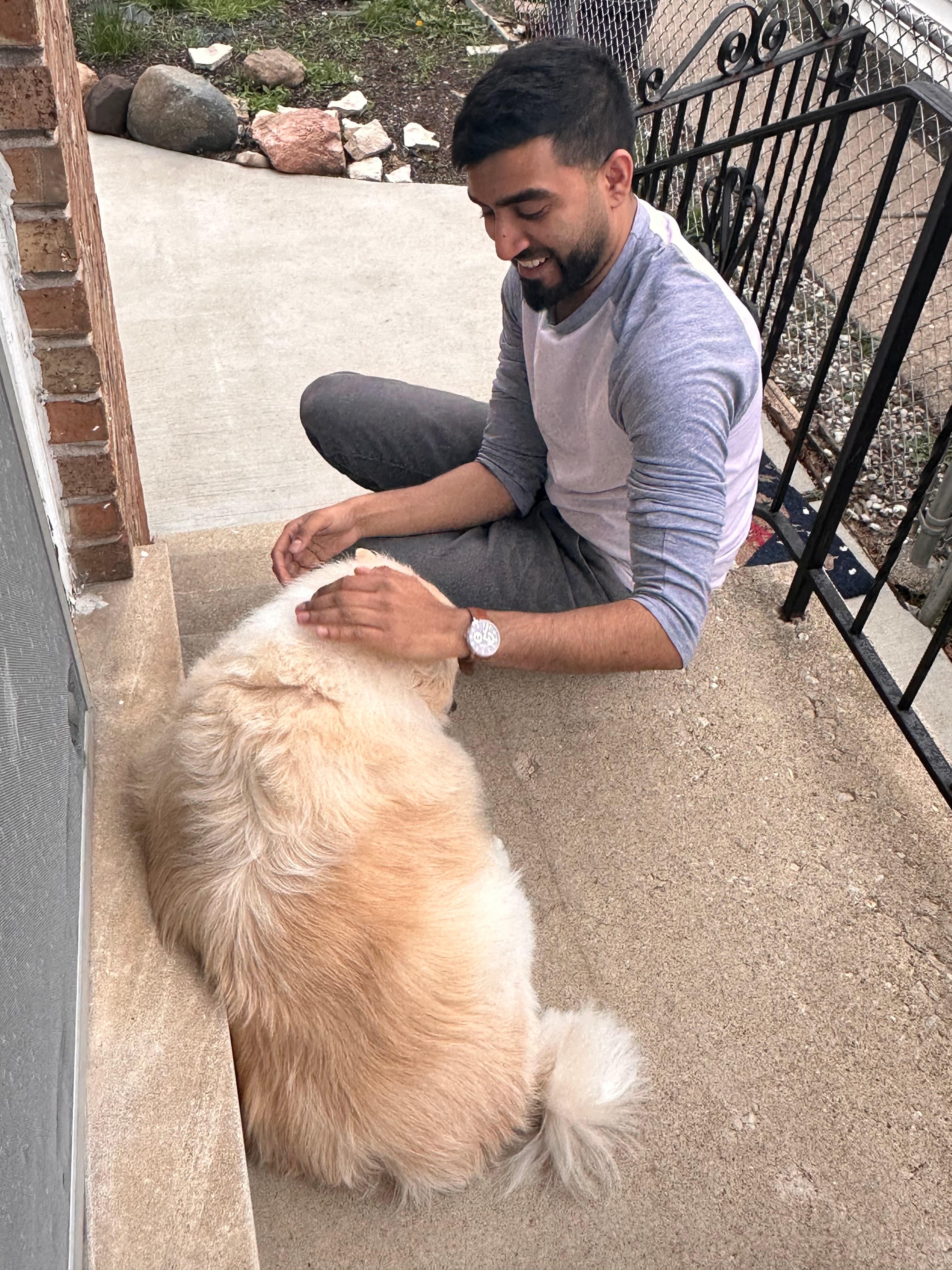 Smiling man in a grey shirt pets a large, fluffy, cream-colored dog outdoors.
