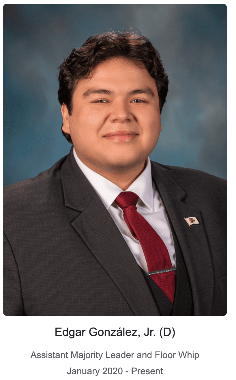 Edgar González, Jr. smiling in a dark suit and red tie against a blue background.