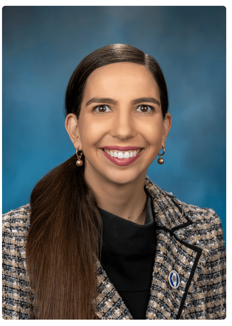 Smiling woman with long brown hair in a side ponytail, wearing a professional tweed blazer.